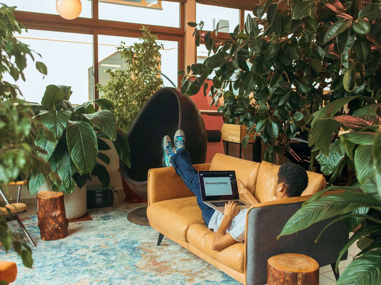 A man reclining on a sofa among houseplants, typing on a laptop showing a marketing strategy document