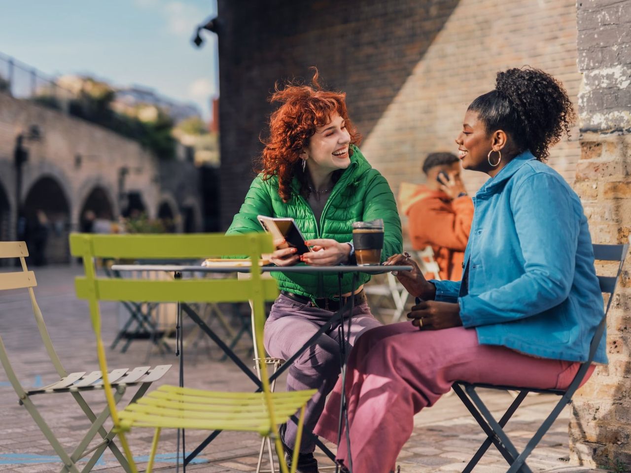 Two women laughing at an outdoor café table, one holding a phone and a coffee