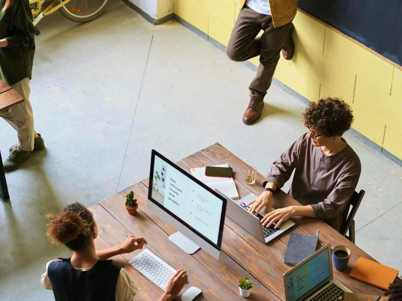 Overhead view of two people working at laptops around a wooden desk, with a colleague standing nearby