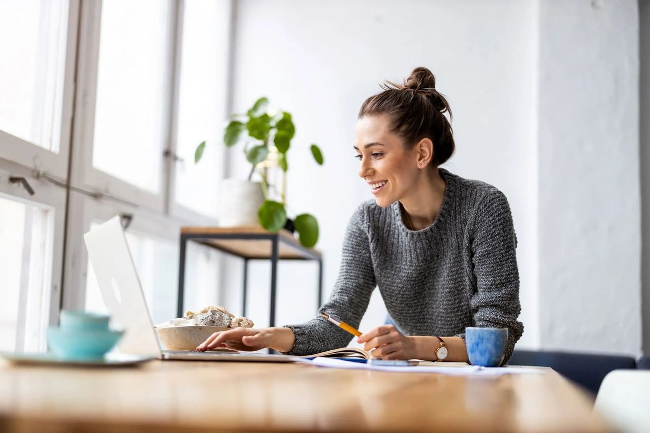 A smiling woman working at a laptop and notebook, with a houseplant and a mug of coffee beside her