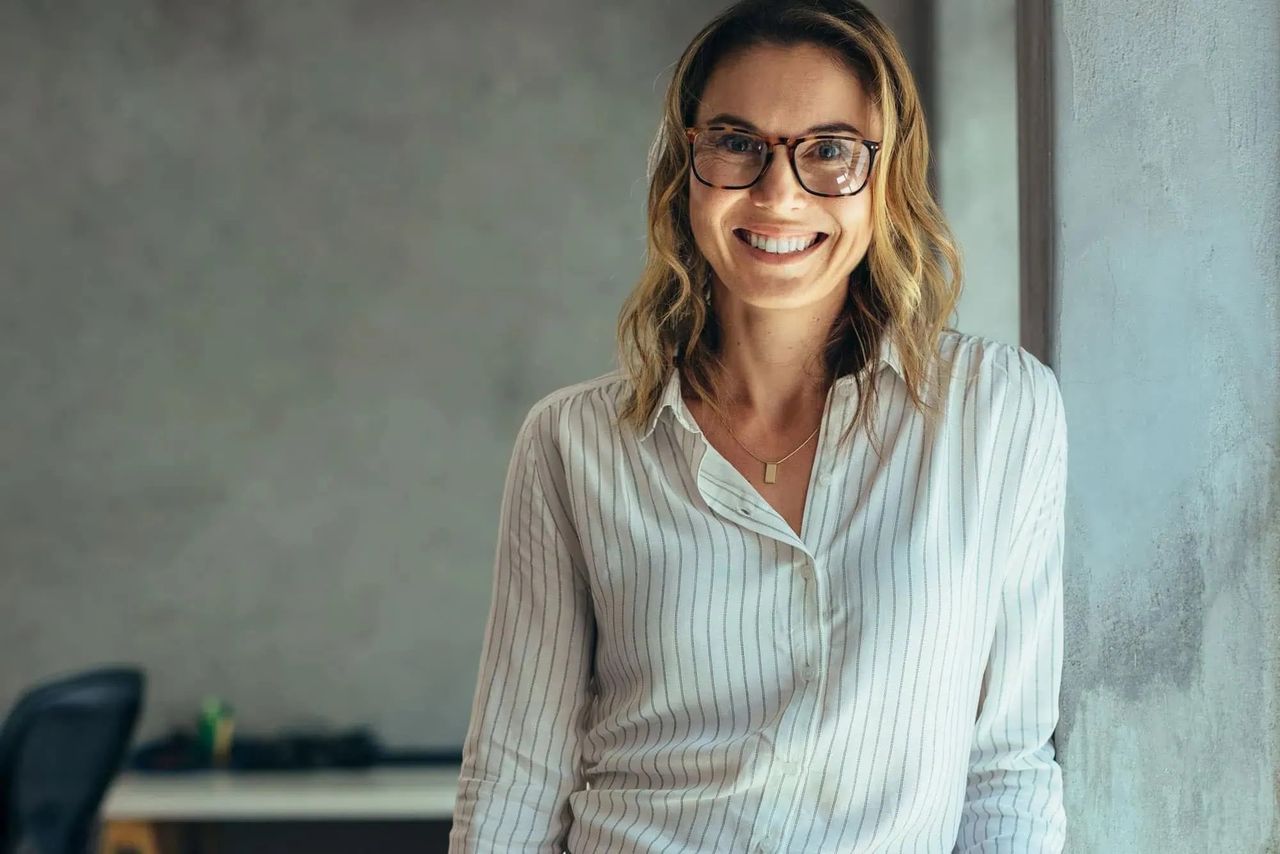 A smiling woman in glasses, standing in an office with a concrete wall behind her