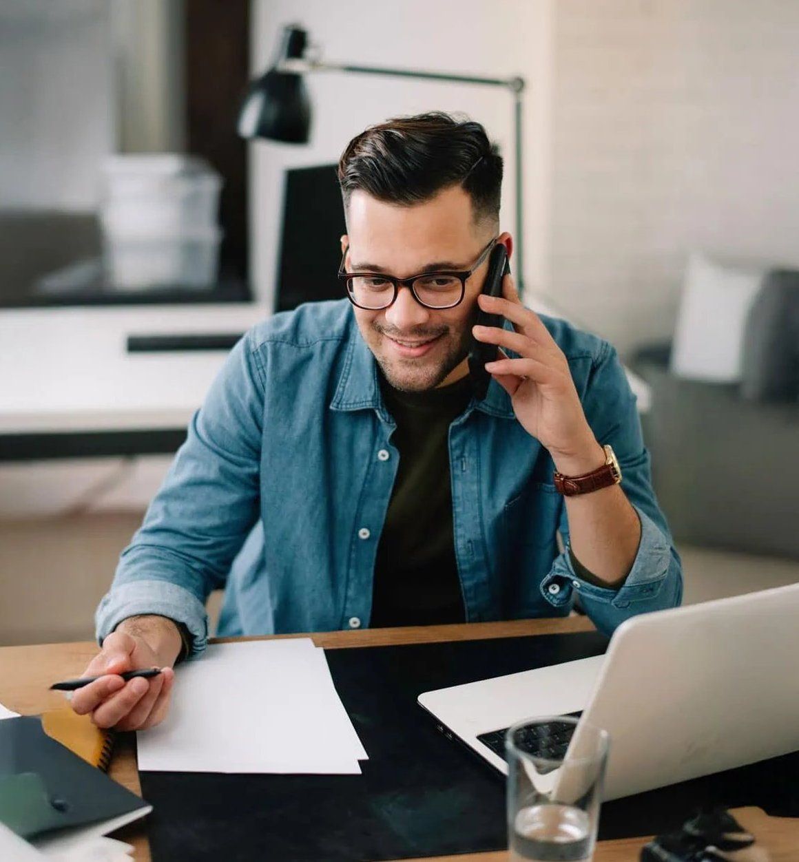 A man in a denim shirt smiling on the phone at his desk, with an open laptop and fabric swatches in front of him