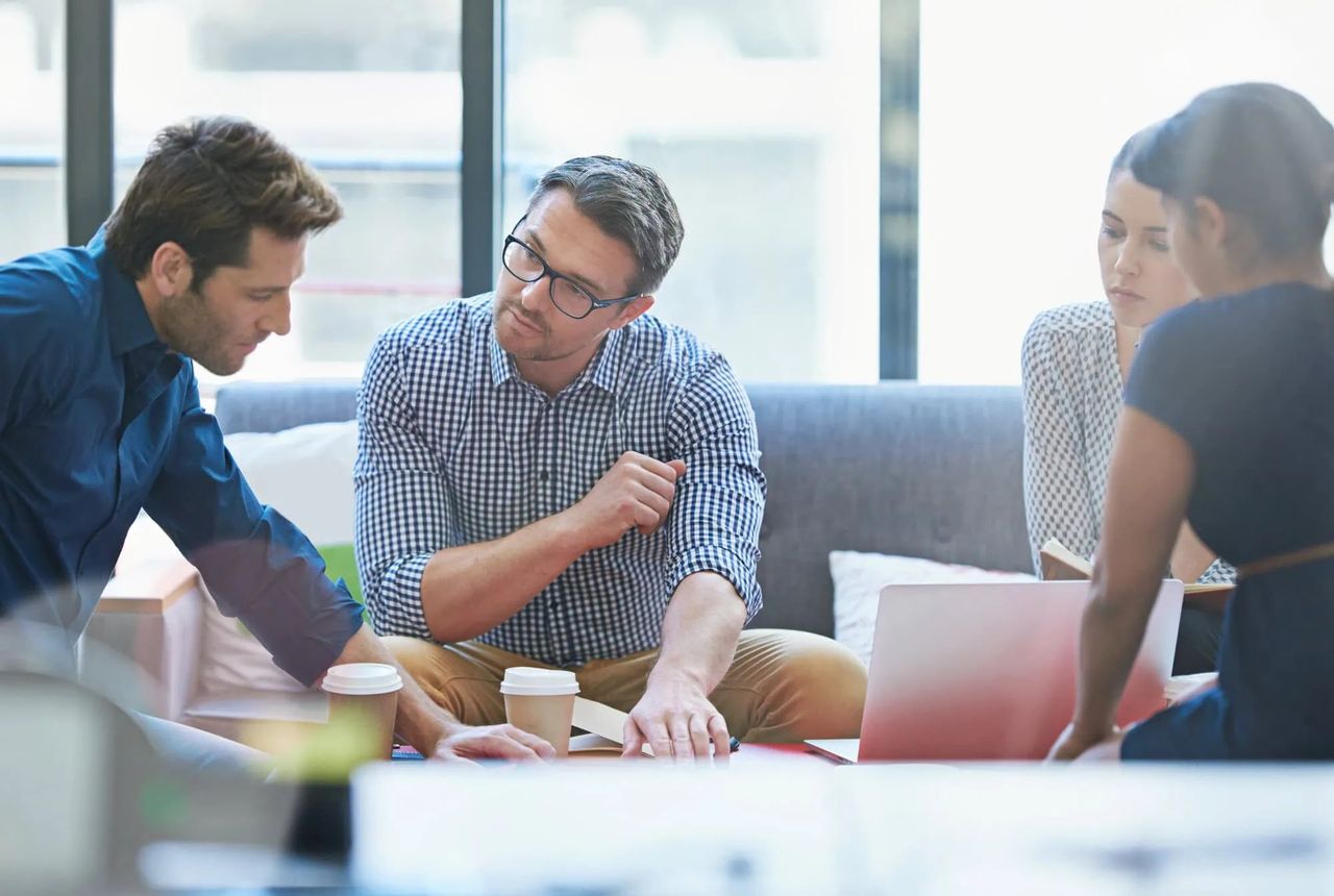 Four colleagues discussing work together in an office, gathered around a laptop with coffee cups on the table
