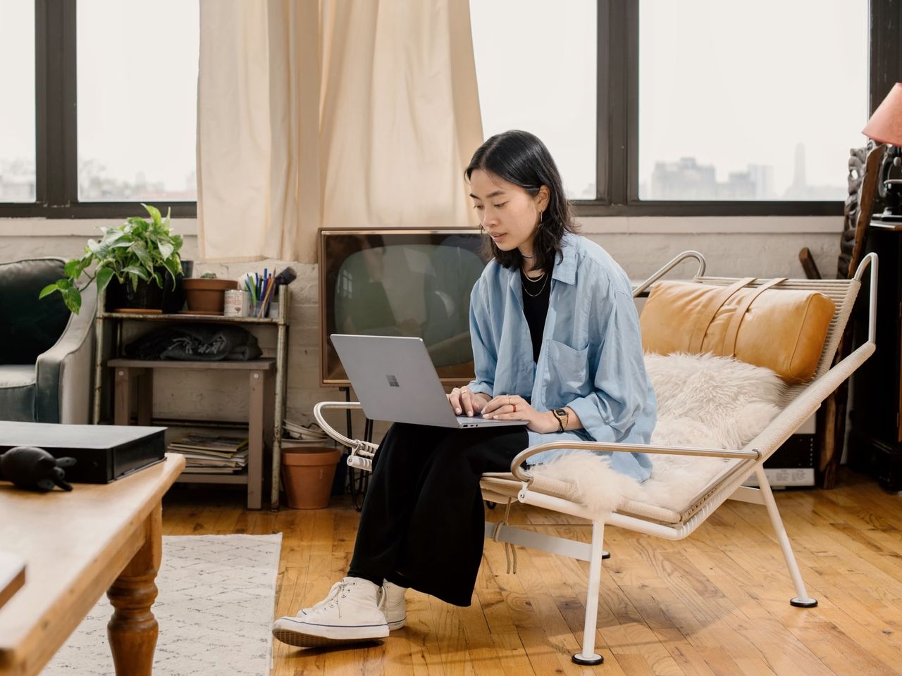 A woman sitting cross-legged on a chair, working on a laptop in a bright, plant-filled apartment
